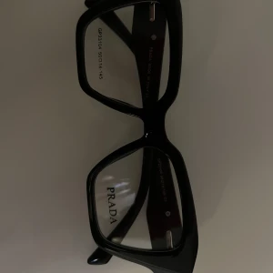 Svarta glasögon från Prada - Snygga svarta glasögon från Prada med markerad, fyrkantig båge och ikonisk logga i silver på sidorna. Glasögonen har en modern och trendig look som verkligen sticker ut. Perfekta för dig som vill ha en stilren accessoar med lyxig känsla. ❗️ALDRIG ANVÄNDA ❗️ splitternya, passar ej min huvudform. Köpta för runt 2000kr. Pris går att diskuteras 