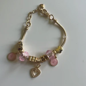 Guldigt armband med rosa berlocker - Charmigt guldarmband med flera berlocker, bland annat rosa glaspärlor, små runda hängen och ett hjärta. Armbandet har en kedja med justerbar längd och detaljer i blank metall. Perfekt accessoar för att lyfta din stil med lite extra färg och bling.