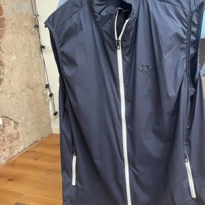 Mörkblå väst från J.Lindeberg - Sportig mörkblå väst från J.Lindeberg med vit dragkedja framtill och på fickorna. Västen har en stilren look med diskret logga på bröstet och är tillverkad i ett lätt syntetmaterial. Perfekt för lager-på-lager och snygg till jeans eller träningsbyxor.