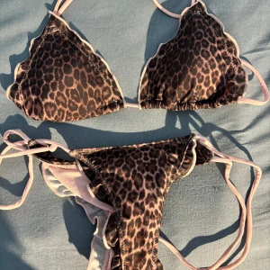 Leopardmönstrad bikini med volangkant - Snygg bikini med leopardmönster och ljusrosa volangkant. Triangeltopp med knyt i nacken och ryggen samt matchande trosa med smala knytband i sidorna. Perfekt för stranden eller poolen. ALDRIG ANVÄND, säljer pga för liten. Toppen är XS och underdelen är S