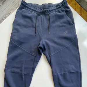 Mörkblå mjukisbyxor från Nike  - Mörkblå mjukisbyxor från Nike med dragsko i midjan och diskret Nike-logga på låret. Byxorna har en smalare passform, ribbad midja och praktiska fickor, varav en med dragkedja bak. Perfekta för chill eller träning.