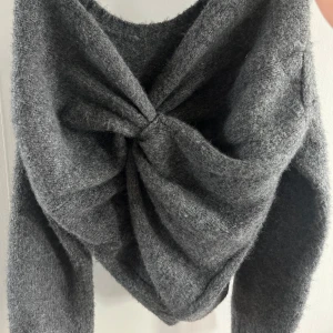 Grå stickad tröja med knutdetalj - Mysig grå stickad tröja med lång ärm och en snygg knutdetalj där bak som ger en cool twist. Tröjan har en lite lös passform och är perfekt för lager-på-lager under kyliga dagar. 