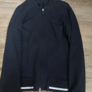 Svart overshirt från Armani Exchange - Svart overshirt från Armani Exchange med dragkedja framtill och diskret logga på bröstet. Jackan har ribbade muddar och vita ränder vid fickorna för en sportig touch. Tillverkad i mjuk bomull, perfekt för lager-på-lager-stil.
