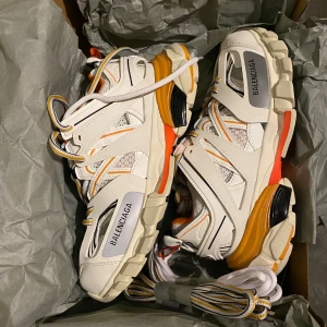 Balenciaga Track sneakers vit/orange 37 - Balenciaga Track sneakers i vitt med orange och gula detaljer, svart och orange sula samt meshpaneler. Märkeslogga på sidan och grov yttersula.  Endast provade inomhus en gång. Två extra skosnören i olika färger från Balenciaga följer med. Säljes pga för små. Nypris över 8000:-. 