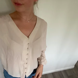 Beige blus med knappar från JDY - Säljer en stilren beige blus från JDY med v-ringning och klädda knappar framtill. Blusen har långa ärmar med lätt puff vid ärmslutet och är gjord i ett tunt, flowy material som ger en luftig känsla. Perfekt att matcha med jeans eller kjol för en clean look.