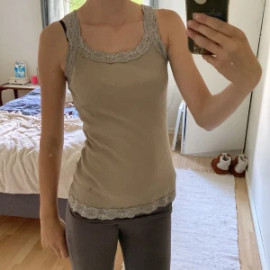 Beige ribbad linnetopp med spets - Supersöt beige ribbad linnetopp med breda axelband och fina spetsdetaljer i ljusgrått längs ringning och nederkant. Tillverkad i 100% bomull, perfekt för en chill och stilren look. Sitter tajt och ärmlös för en enkel men snygg vibe. Osäker på vart den kommer ifrån 
