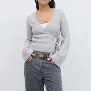 Grå omlottkofta med knyt - Supermjuk grå omlottkofta med knyt i sidan och v-ringning. Koftan är stickad och har långa ärmar med lite utsvängda muddar. Perfekt att slänga på sig över ett linne för en chill och snygg look.