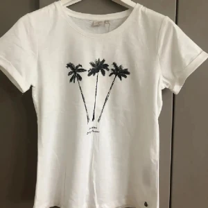 Vit t-shirt med palmer från Cream - Vit t-shirt från Cream med svart palmtryck framtill. Klassisk rund halsringning och korta ärmar. Tillverkad i mjuk bomull, perfekt för en chill och avslappnad stil. Snygg och enkel design som passar till jeans eller shorts.