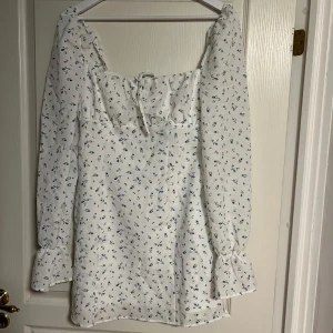 Vit blommig kortklänning med långa ärmar - Supersöt vit kortklänning med små blå blommor, långa puffiga ärmar och volang vid ärmslut. Klänningen har söt snörning vid bysten och är gjord i ett lätt bomullstyg, perfekt för varma dagar. Den har en romantisk och trendig vibe.