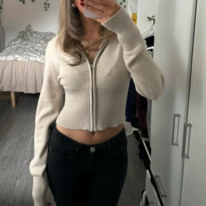 Beige croppad kofta med dragkedja - Superfin croppad kofta i beige med lång ärm och ribbade detaljer. Tröjan har dragkedja framtill och är stickad i mjukt material. Perfekt till jeans eller kjol för en chill och snygg look.