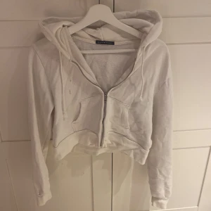 Vit croppad hoodie från Brandy Melville - Säljer en vit croppad hoodie från Brandy Melville med dragkedja framtill och snörning vid huvan. Tröjan har långa ärmar och en stor ficka på magen. Perfekt för chill dagar och enkel att matcha med jeans eller leggings.