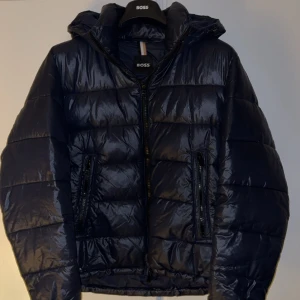 Mörkblå pufferjacka från BOSS - Snygg mörkblå pufferjacka från Hugo Boss med glansig finish och huva. Perfekt för kalla dagar och har en modern, loose passform. Riktigt bra skick knappt använd, storlek S/M. Nypris runt 4000kr