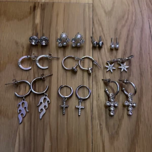 10-pack silverörhängen med hängen - Säljer ett set med 10 par örhängen i silverfärg, blandade stilar med stjärnor, kors, pärlor, flames och glittriga detaljer. Perfekt för dig som gillar att mixa och matcha olika former och uttryck. Alla örhängen är i metall och vissa har strass eller pärlor som dekoration. Vissa örhängen är helt nya och andra kan ha små skavanker men inget som syns på