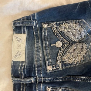 Miss Me jeans med broderade detaljer - Snygga blå jeans från Miss Me med kontrastsömmar och broderade bakfickor med paljetter och silverdetaljer. Jeansen har klassisk femficksdesign, dekorativa knappar och en cool tvättad look. Perfekta för dig som gillar statement-jeans med extra detaljer.