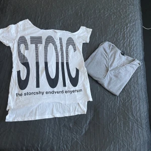 Vit t-shirt med STOIC-tryck plus kofta - Vit t-shirt med stort svart STOIC-tryck på framsidan och coola prickdetaljer i bokstäverna. Modellen är kortärmad med rund hals och har en loose passform. Materialet känns som mjuk bomull, perfekt för chill dagar. Snygg statement-tee för dig som gillar grafiska prints.plus kofta i samma storlek 