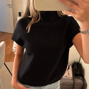 Svart polotopp i stickad material - Säljer en svart polotopp med korta ärmar i mjukt stickat material. Toppen har en hög krage och en loose passform som ger en chill vibe. Perfekt att matcha med jeans eller kjol för en enkel och snygg look.