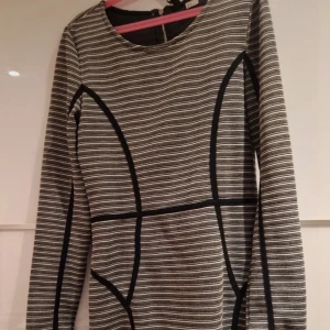Randig grå och svart klänning - Snygg kortklänning med horisontella ränder i grått och svart, samt svarta markerade sömmar som ger en cool grafisk look. Klänningen har lång ärm och rund halsringning, perfekt för dig som gillar stilrena och moderna detaljer.