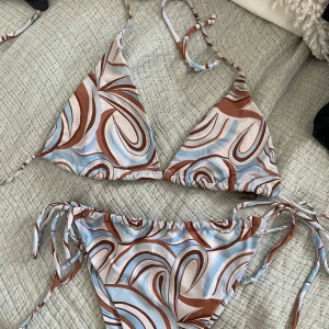 Bikini med snörning - Säljer en snygg bikini med swirlmönster i brunt, ljusblått och vitt. Toppen är trekantsmodell med knyt i nacken och ryggen, och trosan har snörning i sidorna. 