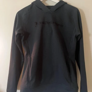 Blå  hoodie Peak Performance - Mörk blå hoodie från Peak Performance med broderad logga på bröstet. Tröjan har huva, långa ärmar och en clean, slim passform. Materialet är mjukt och känns som bomull blandat med polyester, perfekt för chill eller träning. Snygg och enkel design utan extra detaljer.