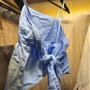 Ljusblå linnetopp med knyt - Supersöt ljusblå linnetopp med smala axelband och volang nedtill. Toppen har en stor knytning framtill som ger en extra touch och sitter snyggt med en lätt croppad passform. Perfekt för varma dagar och chill vibes.