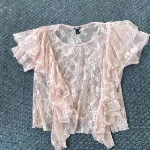 Ljusrosa spetsblus med volanger - Säljer en ljusrosa blus i transparent spets med blommigt mönster. Blusen har korta ärmar och snygga volangdetaljer längs sidorna och axlarna. Perfekt för dig som gillar romantisk och feminin stil. Supersnygg att styla med ett linne eller bralette under.