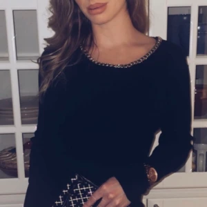 Svart långklänning med kedjedetalj - Svart långklänning från Axisu med långärmad design och en snygg guldfärgad kedja runt halsringningen. Klänningen har en slim passform och en liten slits framtill. Perfekt för dig som gillar stilrena och coola detaljer.