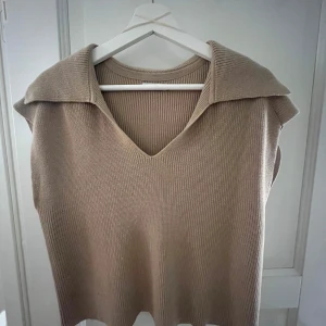 Beige stickad väst från Stockh LM - Säljer en beige stickad väst från Stockh LM med bred krage och v-ringning. Västen har en ribbad struktur och är ärmlös, vilket gör den enkel att styla över en skjorta eller t-shirt. Perfekt för lager-på-lager och snygg till både jeans och kjol.