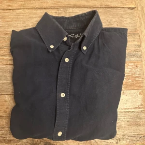 Mörkblå skjorta från Baracuta - Snygg mörkblå skjorta från Baracuta med klassisk button-down krage och vita knappar. Skjortan har en bröstficka med diskret broderad logga och är tillverkad i mjuk bomull. Perfekt för dig som gillar stilrena och tidlösa plagg. Köpt från MQ