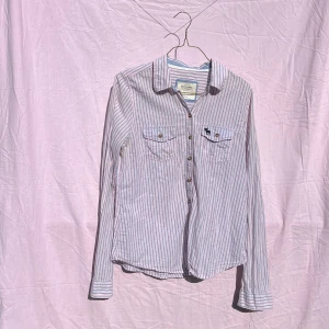 Randig skjorta från Abercrombie & Fitch - Snygg skjorta i ljusrosa och ljusblå ränder från Abercrombie & Fitch. Modellen har klassisk krage, två bröstfickor med knappar och ett litet broderat logo-djur på ena fickan. Tillverkad i 100% bomull, perfekt för dig som gillar en chill och stilren look.