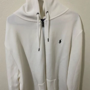 Vit hoodie från Polo Ralph Lauren - Säljer en fräsch vit hoodie från Polo Ralph Lauren med dragkedja och snörning vid huvan. Tröjan har det klassiska Polo-logot broderat på bröstet och praktiska fickor framtill. Perfekt för chill dagar och enkel att matcha med jeans eller joggers.