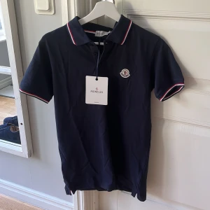 Mörkblå Moncler piké t-shirt M - Snygg mörkblå piké t-shirt från Moncler med klassisk krage och korta ärmar. T-shirten har vita och röda ränder på krage och ärmslut samt Moncler-logga på bröstet. Tillverkad i mjuk bomull som känns skön mot huden. Perfekt för dig som gillar stilrena märkesplagg. Väldigt bra skick.