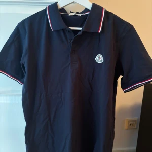 Marinblå Moncler piké oanvänd ny - Snygg marinblå pikétröja från Moncler i storlek L. Klassisk krage med röd, vit och blå kant, korta ärmar och Moncler-logga på bröstet. Tröjan har knappar framtill och är gjord i mjuk bomull som känns skön mot huden. Perfekt för dig som gillar stilrena och sportiga plagg. Helt oanvänd och ny.