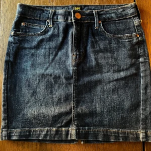 Mörkblå jeanskjol från Lee - Snygg mörkblå jeanskjol från Lee med klassisk femficksdesign, slitstark denim och guldfärgade detaljer. Kjolen har dragkedja och knapp framtill, samt Lee-lapp bak i brunt. Perfekt för dig som gillar en chill och tidlös look.