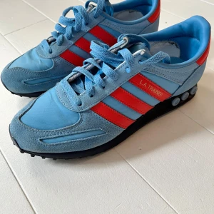 Adidas L.A. Trainer blå/röda sneakers  - Säljer ett par Adidas L.A. Trainer sneakers i ljusblå mocka och textil med röda detaljer och klassiska tre ränder på sidan. Skorna har snörning, svart  sula med dämpningspluggar och rund tå. Perfekt för dig som gillar retrostil och färgkombon blå/röd. Nypris 950 kr, gär att förhandla pris!