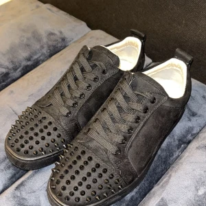 Svarta Loubs  - Säljer ett par svarta sneakers från Christian Louboutin med coola svarta nitar på tån och klassisk röd sula. Skorna är i mocka med snörning och har en rund tå. Kommer med originalkartong och dustbag. Perfekt för dig som gillar edgy designerstil.