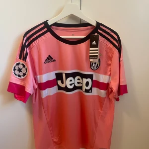 Juventus Pogba rosa fotbollströja M - Juventus fotbollströja med Pogba 10 på ryggen, tillverkad av lätt och ventilerande polyester. Tröjan är rosa med svarta och vita detaljer, har svarta Adidas-ränder på axlarna och Champions League-märke på ärmen. Jeep-logga på bröstet och klubbmärke med stjärna.
