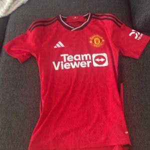 Manchester United röd matchtröja Adidas S - Säljer en officiell Manchester United matchtröja från Adidas i storlek S. Tröjan är röd med svarta detaljer, vit TeamViewer-logga och klubbmärke på bröstet. Den har korta ärmar, sportig passform och är gjord i lätt polyester. Snygga mönster i tyget och klassisk krage med ränder.