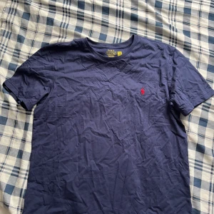 Marinblå t-shirt från Polo Ralph Lauren - Snygg marinblå t-shirt från Polo Ralph Lauren med klassisk röd broderad logga på bröstet. Modellen är kortärmad och har en rund hals. Tillverkad i mjuk bomull som är skön mot huden. Perfekt för dig som gillar stilrena och ikoniska plagg.