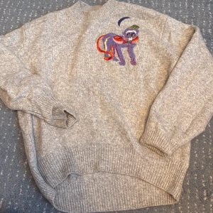 Grå stickad tröja med broderad apa - Grå stickad tröja från H&M i storlek XS med en färgglad broderad apa på bröstet. Apan har lila, orange och gröna detaljer samt pärlor som ger extra bling. Tröjan har ribbade muddar och lång ärm, perfekt för dig som gillar unika detaljer.