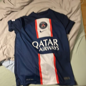 PSG Mbappé 7 Nike fotbollströja M - Säljer en officiell Paris Saint-Germain fotbollströja med Mbappé 7 på ryggen. Tröjan är marinblå med vit och röd vertikal panel på framsidan, PSG-logga och Qatar Airways tryck. Tillverkad i Dri-Fit material som andas, perfekt för match eller träning.