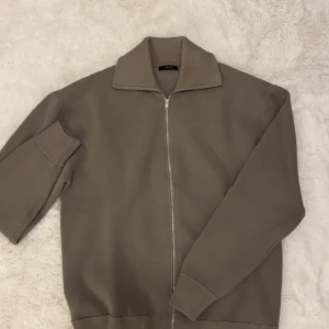 Oliv Grön stickad kofta från Massimo Dutti - Säljer en oliv grön stickad kofta från Massimo Dutti med dragkedja framtill och hög krage. Tröjan har långa ärmar med breda muddar och är perfekt för lager-på-lager. Enkel och stilren design som funkar till många olika looks.