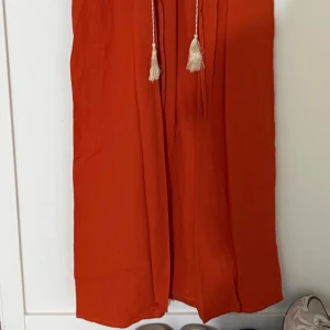 Broderad orange klänning från SHEIN - Superfin orange långklänning från SHEIN med broderade detaljer i vitt, blått och gult framtill och på ärmarna. Klänningen har v-ringning, snörning med tofsar och långa ärmar. Perfekt för dig som gillar boho-stil och färg!