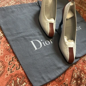 Vita och bruna pumps från Dior - Snygga pumps från Dior i vitt skinn med bruna detaljer framtill och bak. Skorna har spetsig tå, hög klack och dekorativa perforeringar längs kanterna. Perfekt för dig som gillar klassisk stil med en twist. CD-logga i guld på insidan.
