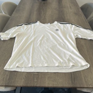 Vit Adidas t-shirt med svarta ränder - Vit t-shirt från Adidas med klassiska svarta ränder längs axlarna och det ikoniska treklöver-loggan på bröstet. Modellen är kortärmad och har en rund halsringning. Skön passform och tillverkad i mjuk bomull, perfekt för en chill och sportig stil.