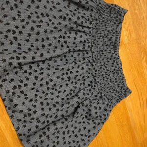 Blå blommig kortkjol med smock - Supersöt grå kortkjol med svart blommigt mönster och bred smock i midjan. Kjolen har en lätt volangig form och är gjord i ett mjukt, ribbat material som känns skönt mot huden. Perfekt för dig som gillar en chill och trendig look. Skriv om ni har frågor!