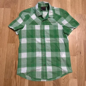 Grön och vit rutig Mammut skjorta - Storlek: S Snygg kortärmad skjorta från Mammut i grönt och vitt rutigt mönster. Skjortan har klassisk krage, knappar framtill och är tillverkad i en skön blandning av bomull och polyester. Perfekt för dig som gillar friluftsliv eller vill ha en chill stil.