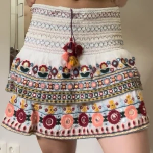Broderad vit, kort volangkjol - Superfin vit kortkjol med broderade blommor i rött, rosa, gult och grönt. Kjolen har bred smock i midjan, snörning med tofsar och volangkant. Materialet känns som bomull och kjolen har färgglada detaljer som gör den unik och boho.