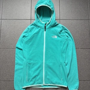 Turkos fleecejacka från The North Face - Storlek: S Turkos fleecejacka från The North Face med huva och hel dragkedja framtill. Jackan har två sidofickor med dragkedja och är tillverkad i mjuk polyester. Perfekt för kyliga dagar och outdoor-häng. Regular fit och långärmad design.