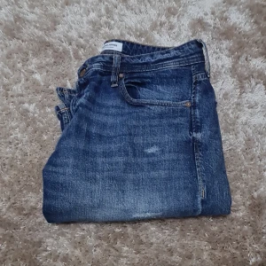Blå jeans från Jack & Jones - Säljer ett par klassiska blå jeans från Jack & Jones med snyggt slitna detaljer och fem fickor. Jeansen har normal passform och raka ben, med en cool tvättad look och kontrastsömmar. Perfekt för dig som gillar en avslappnad stil.