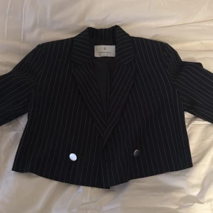 Svart randig croppad kavaj Stradivarius - Snygg svart croppad kavaj från Stradivarius med vita smala ränder och två silvriga knappar framtill. Klassisk krage och lång ärm, perfekt för att ge din outfit en cool edge. Stilren och trendig design som sticker ut.